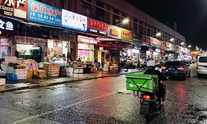 周二，上海建阳水产品批发市场沿街，一名送货快递员骑着摩托车。每一个进入市场的人都要出示健康码并戴上口罩，而运输货物进入市场的商户则要出示相应的证明。